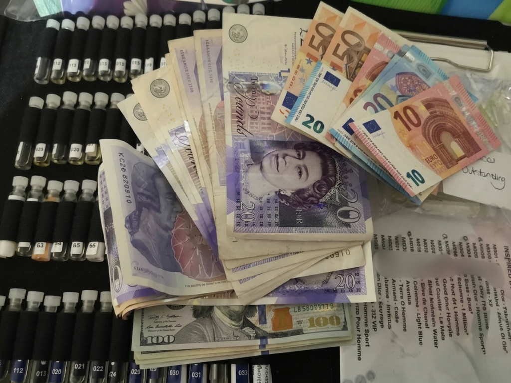 A collection of perfume sample vials arranged neatly next to a fan of international currency, including British Twenty Pound notes, US One Hundred Dollar bills, and various Euro notes.